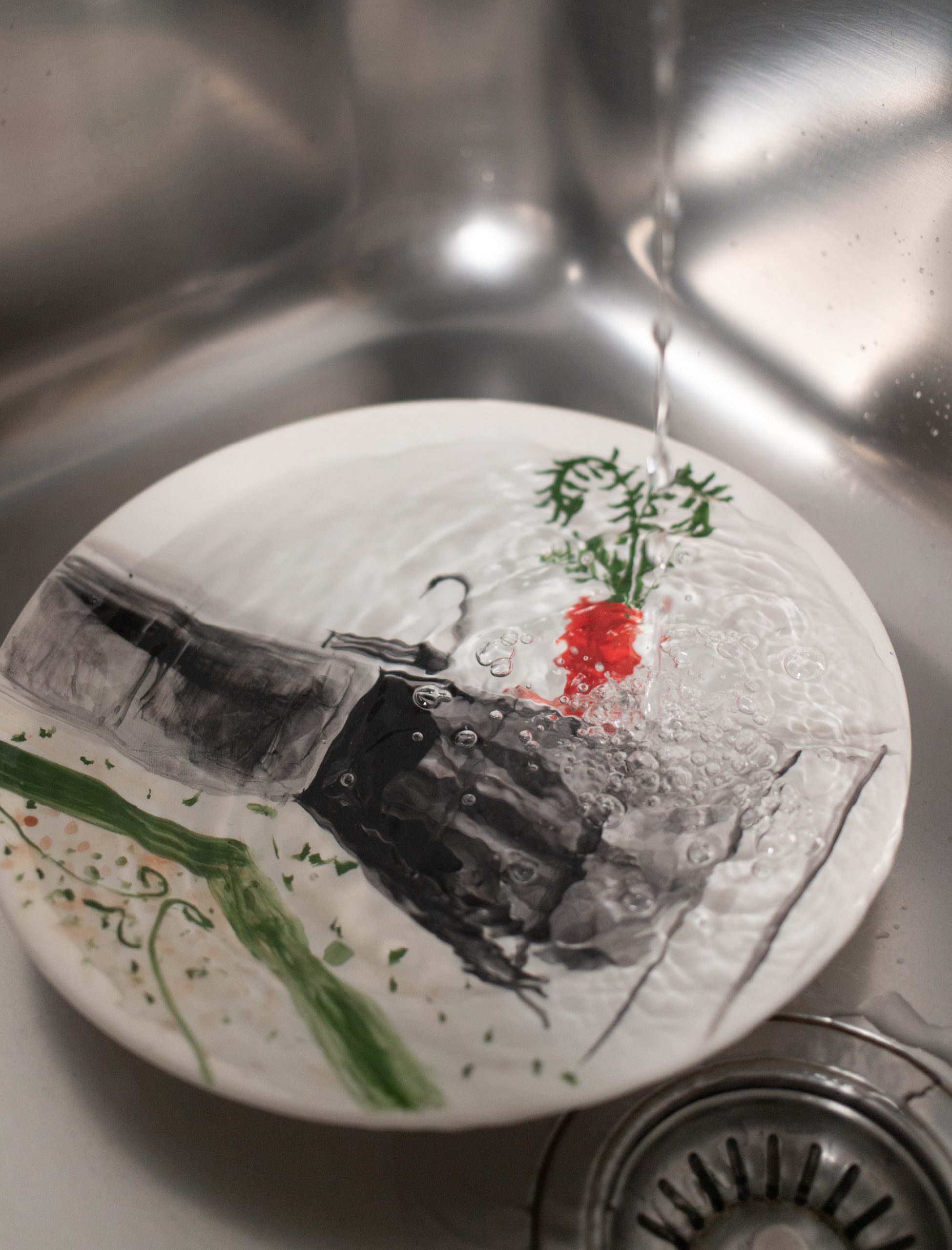 “Dettaglio di un piatto illustrato sotto acqua corrente, con decorazioni artistiche astratte e vegetali visibili attraverso il movimento dell’acqua.