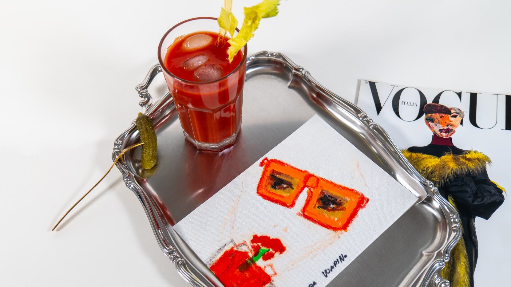 Vassoio in metallo con un bicchiere di cocktail rosso, un cetriolino e una cartolina illustrata; accanto, una rivista di moda su sfondo bianco.