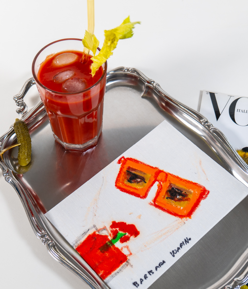 Vassoio in metallo con un bicchiere di cocktail rosso, un cetriolino e una cartolina illustrata; accanto, una rivista di moda su sfondo bianco.