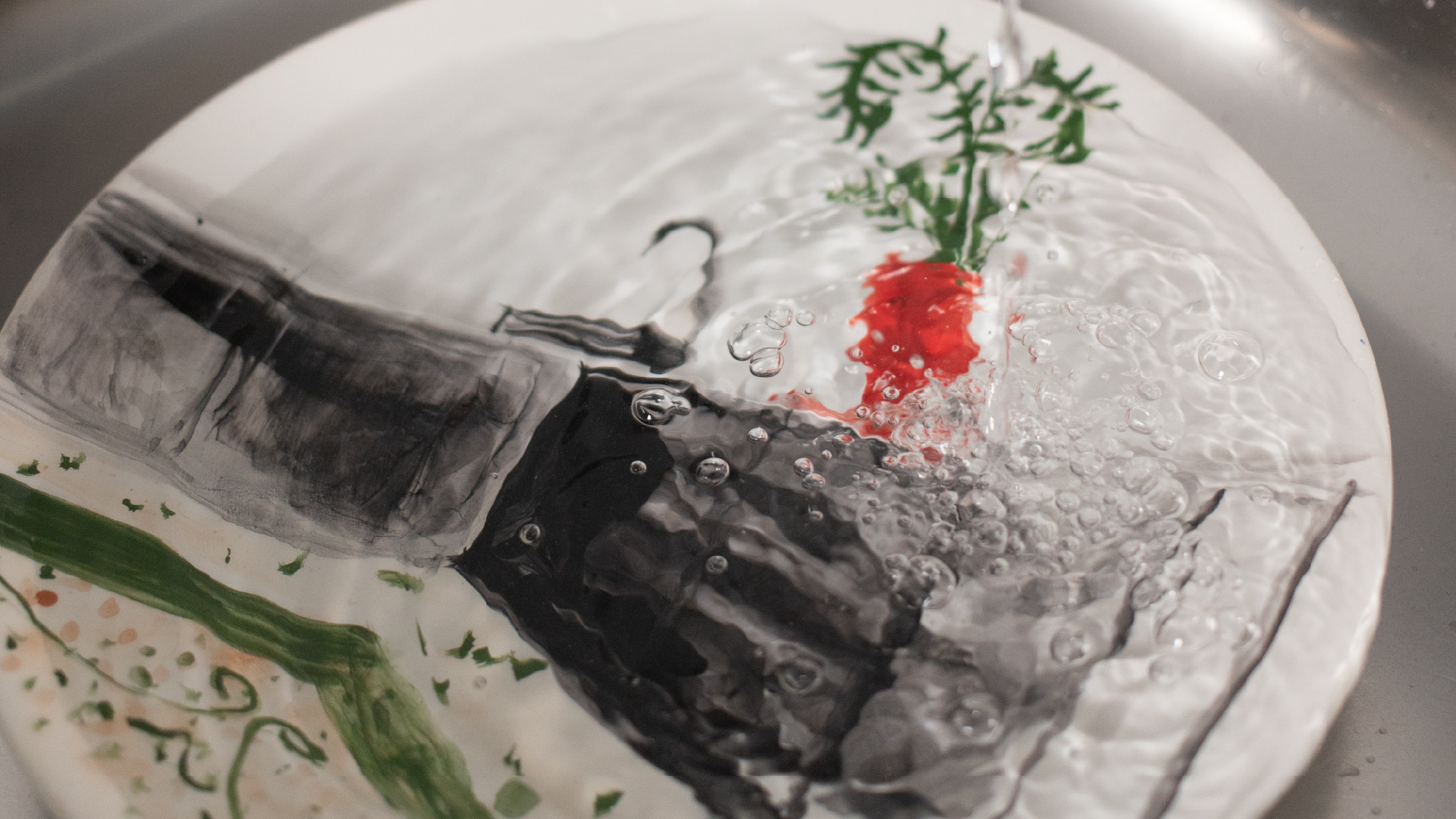 “Dettaglio di un piatto illustrato sotto acqua corrente, con decorazioni artistiche astratte e vegetali visibili attraverso il movimento dell’acqua.
