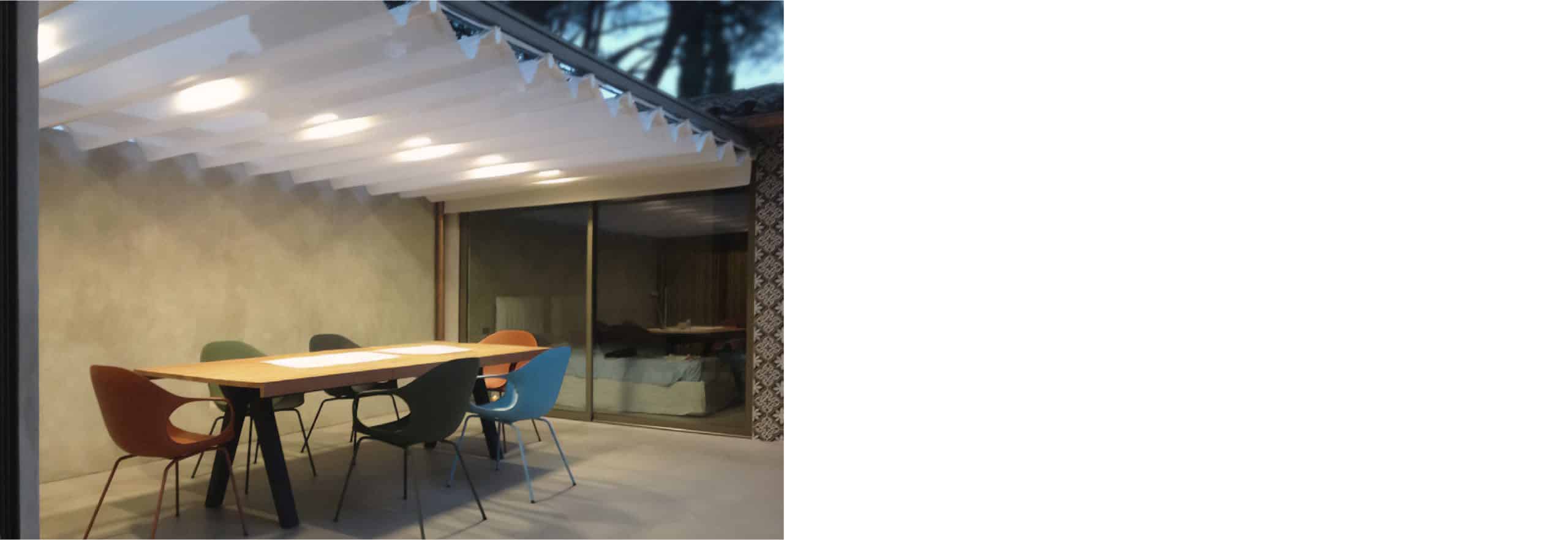 Terrazza coperta con tavolo in legno, sedute colorate e illuminazione integrata dal design contemporaneo.