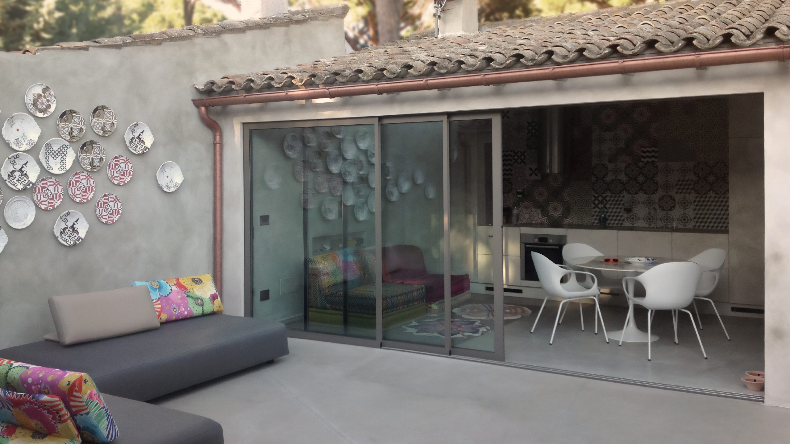 Vista del patio esterno con divano grigio, cuscini colorati, parete decorata e cucina interna visibile attraverso la vetrata scorrevole.