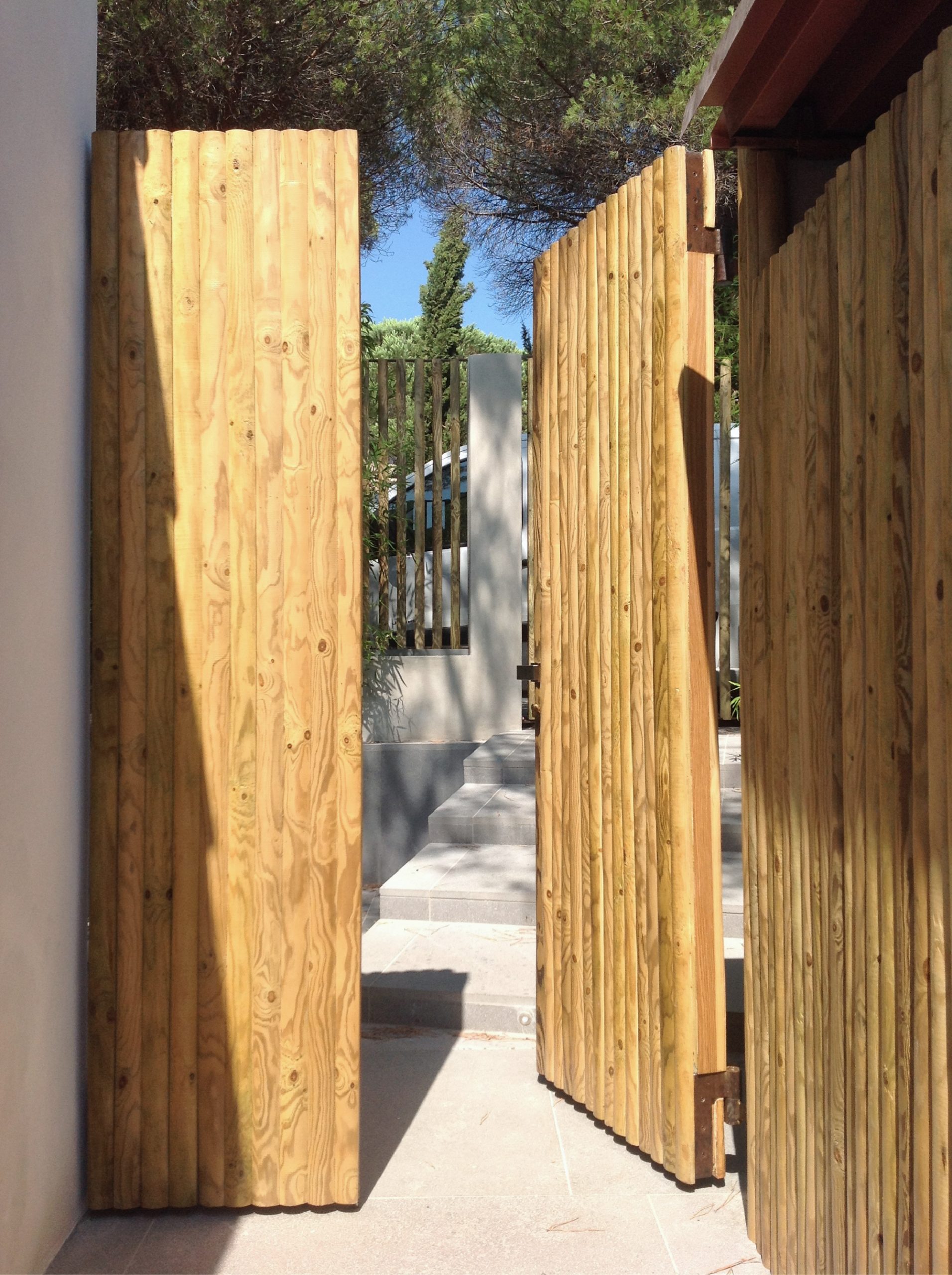Ingresso esterno con cancelli in legno naturale aperti, pavimentazione chiara e vegetazione mediterranea intorno al bastidon.