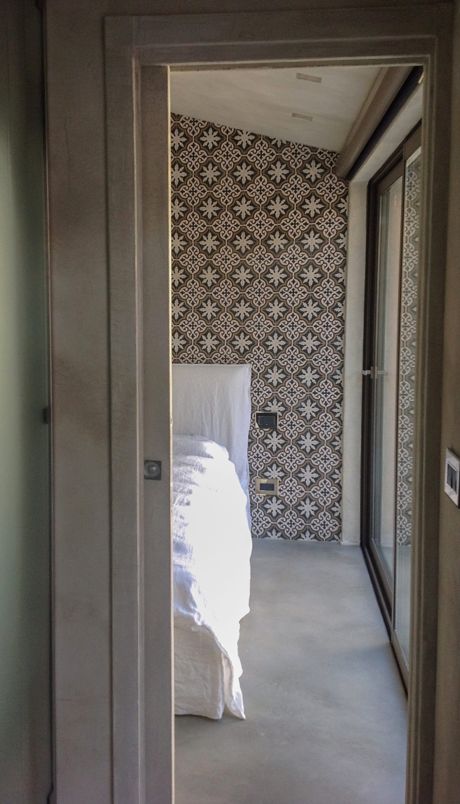 Vista dalla porta verso la camera da letto del bastidon, con parete decorativa a piastrelle, letto bianco e vetrata laterale.