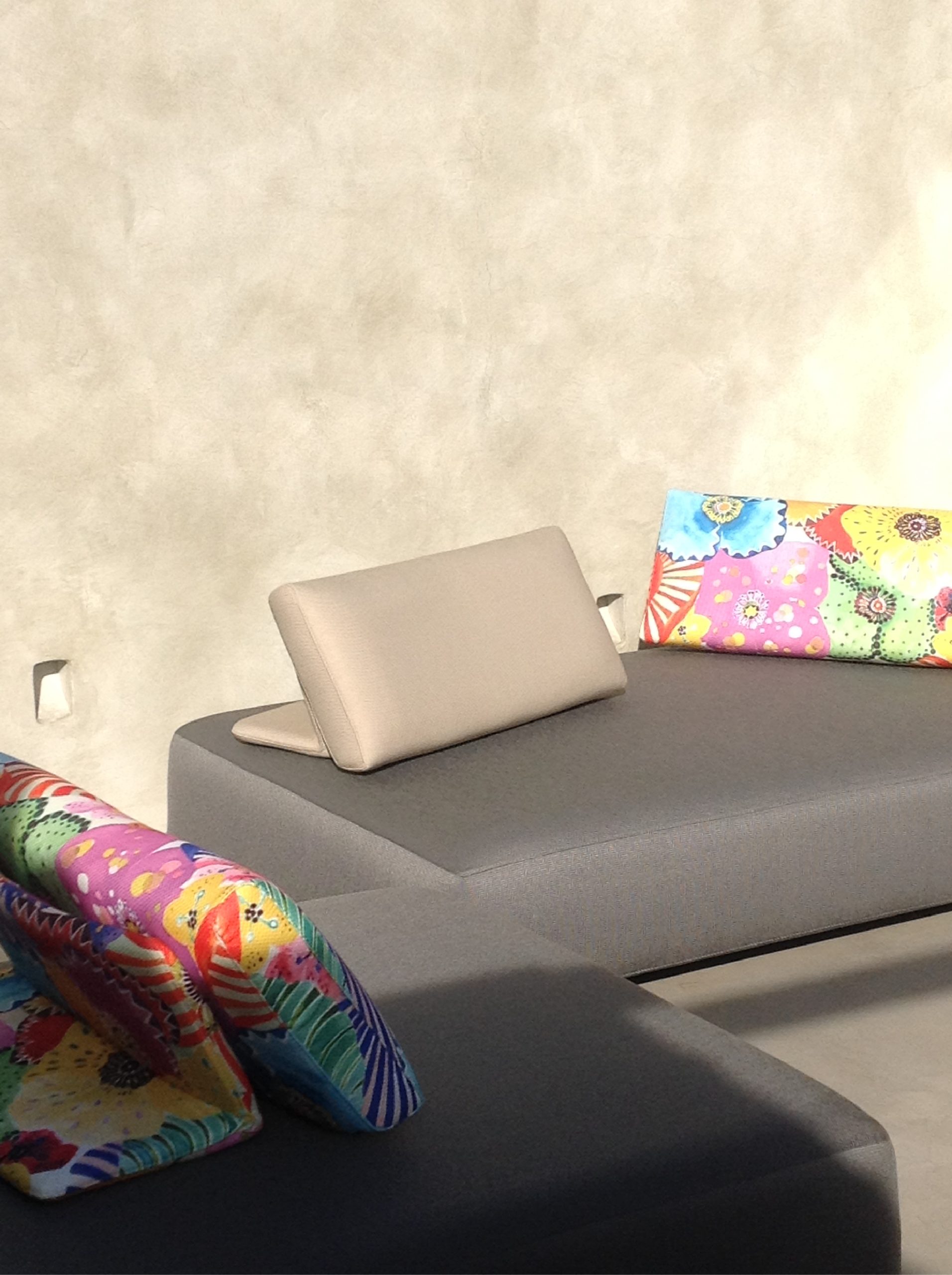 Area relax esterna con divano grigio, cuscini colorati e parete materica chiara nel progetto A Bastidon in Saint-Tropez.