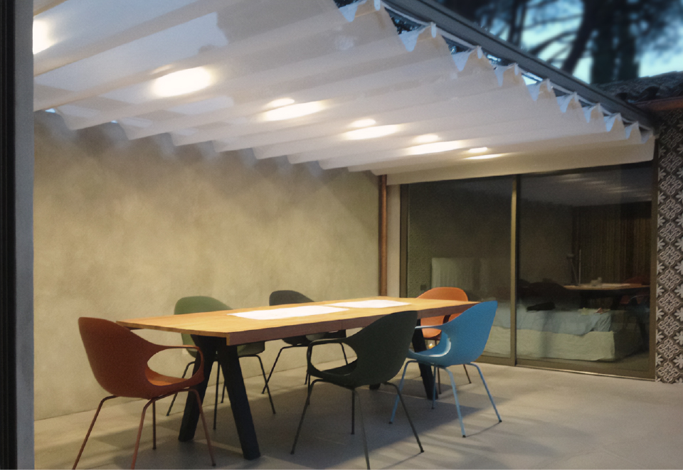 Zona pranzo esterna serale con pergola bianca illuminata, tavolo in legno e sedie colorate nel patio del bastidon